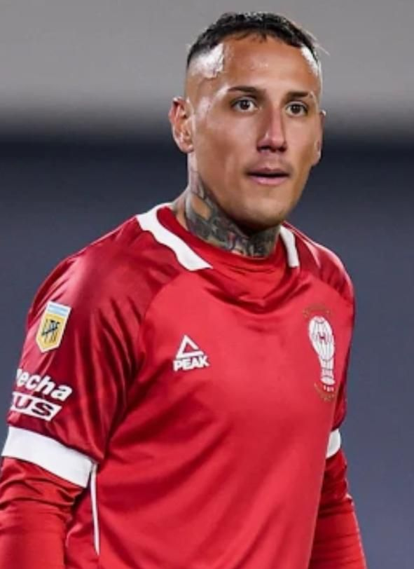 Jugador de fútbol con camiseta roja, tatuajes en el cuello, mirando hacia adelante.