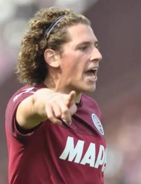 Jugador de fútbol con camiseta granate y pelo rizado, apuntando hacia delante con la boca abierta.