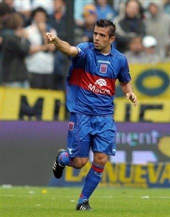 Un jugador de fútbol con uniforme azul celebra un gol, con el puño levantado, corriendo en un campo verde.