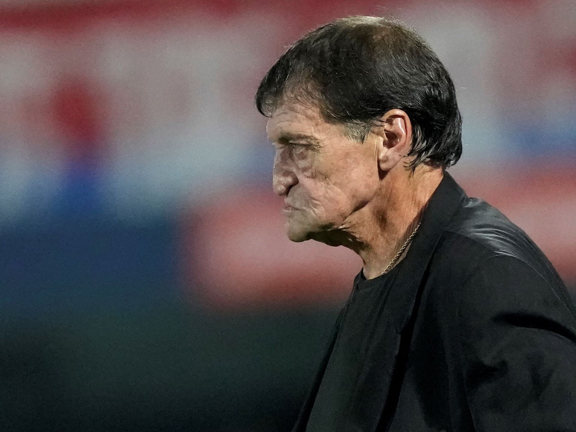 Entrenador de fútbol con cabello oscuro, expresión seria y camiseta negra. Fondo del estadio.