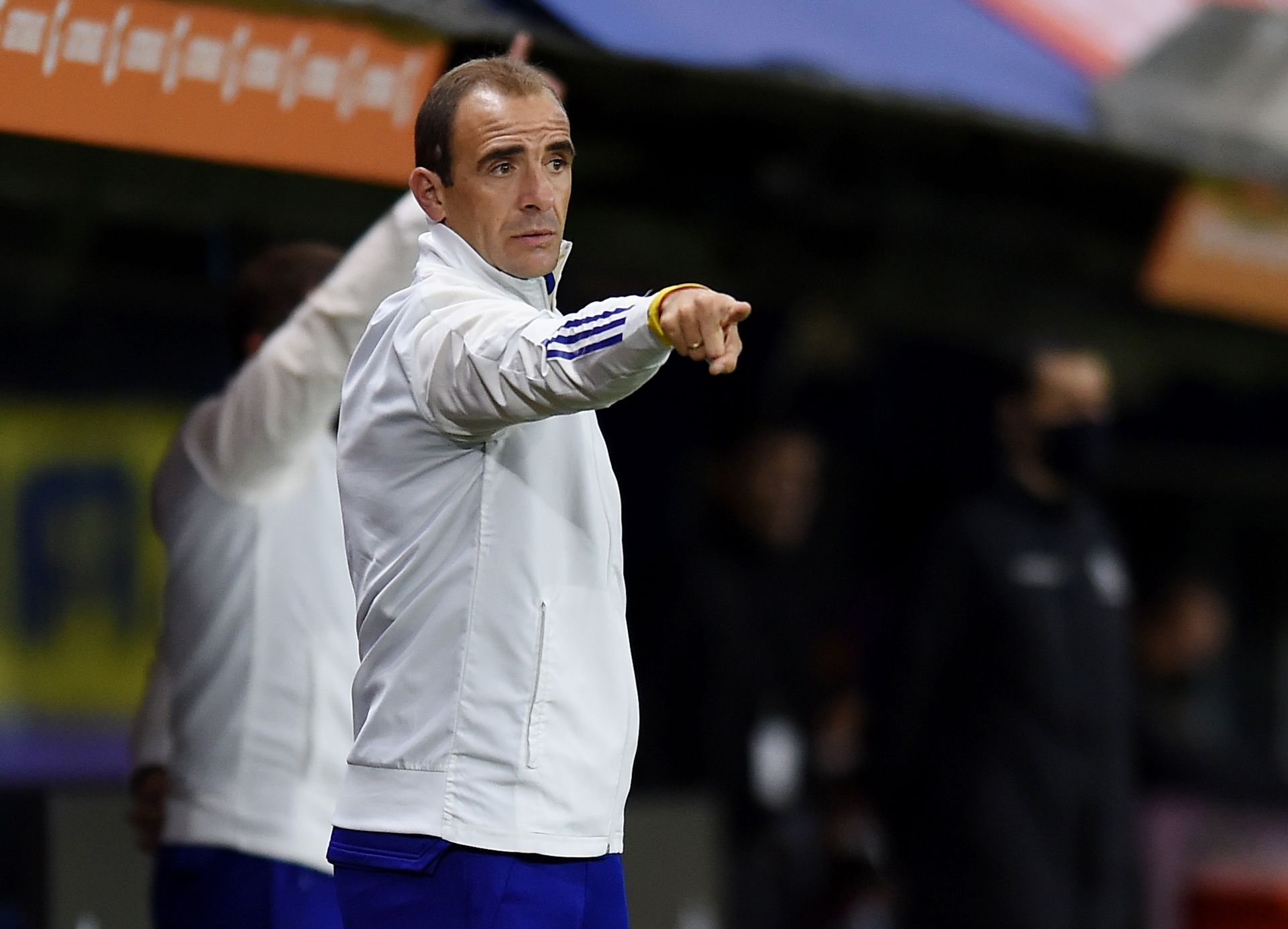 Entrenador de fútbol señalando, vistiendo chaqueta blanca, mirando el juego en el campo.