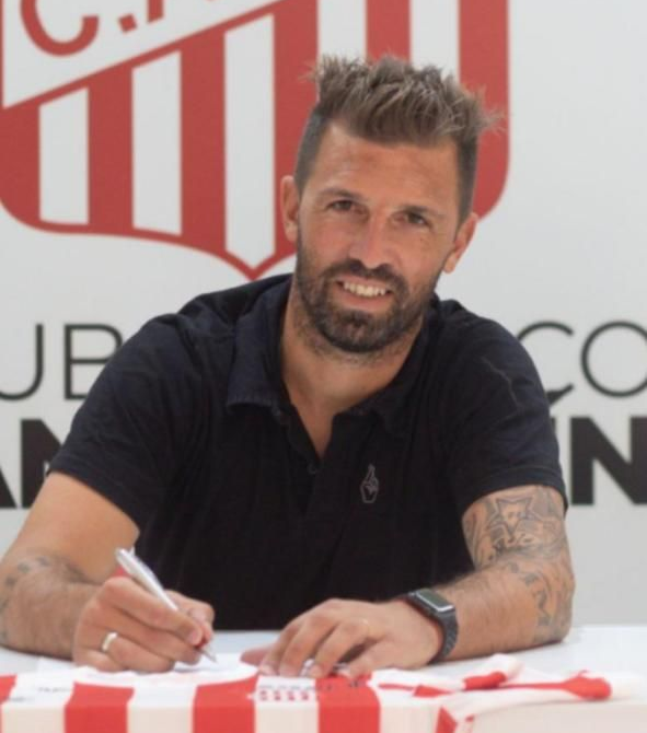 Hombre con camisa negra firmando un documento, sonriendo. El logo del club al fondo.
