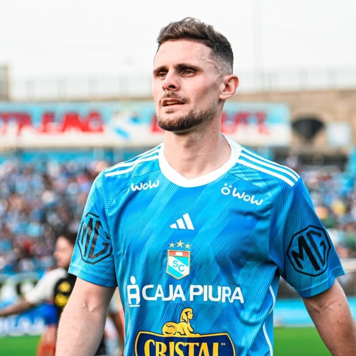 Jugador de fútbol con una camiseta azul claro, mirando hacia arriba con una expresión seria en el campo.
