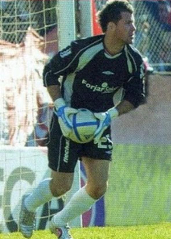 Un hombre con polo azul marino sonríe, levanta el pulgar y firma un documento con el logo del club de fondo.