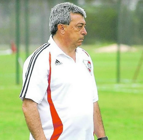 Entrenador de fútbol con camiseta negra mirando hacia abajo, vista de perfil, fondo del estadio.