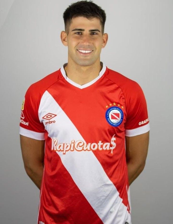 Jugador de fútbol con una camiseta roja y blanca, sonriendo, de pie frente a un fondo blanco.