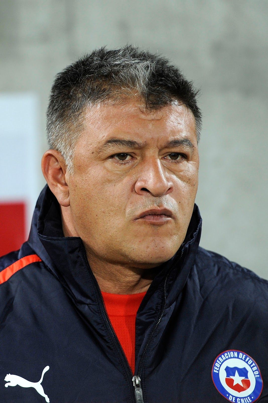 Manuel Pellegrini, entrenador de fútbol chileno, vistiendo una chaqueta azul con una camisa roja debajo.