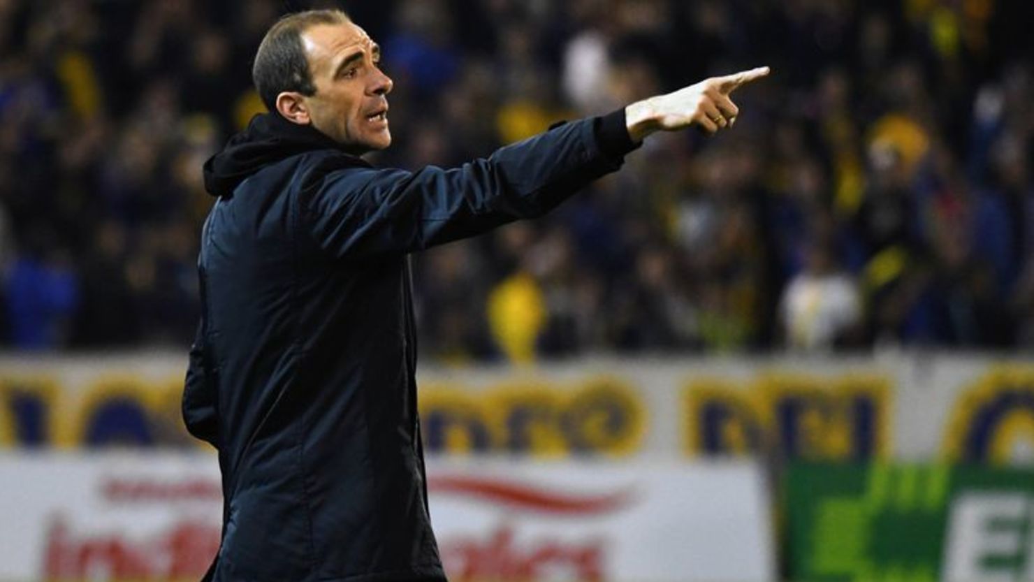 Entrenador de fútbol apuntando hacia el campo, vistiendo un abrigo oscuro, con una expresión concentrada, en un estadio.