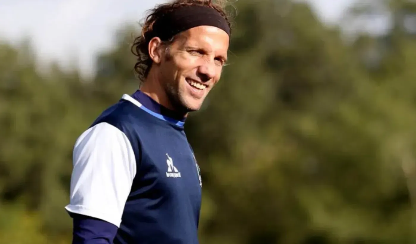 Jugador de fútbol sonriendo, vistiendo una diadema oscura y una camiseta deportiva azul al aire libre.