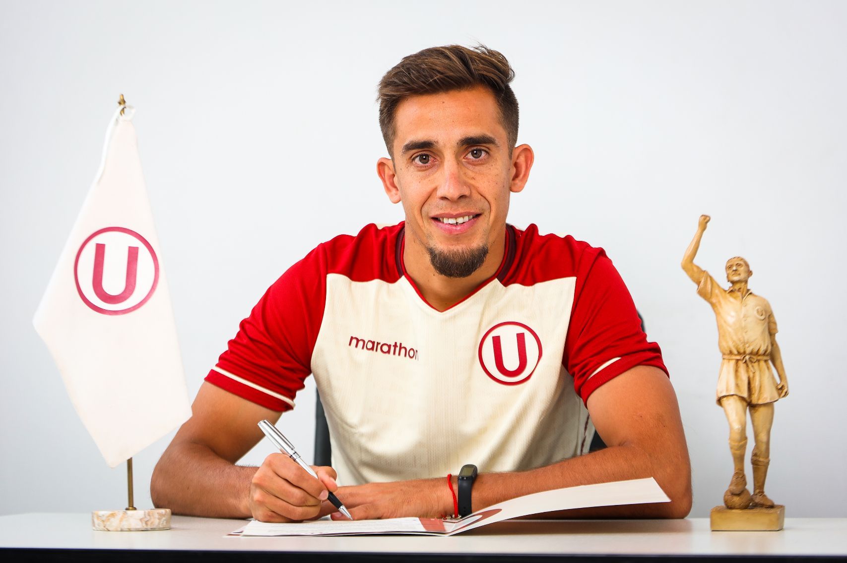 Jugador de fútbol firmando un documento en una mesa, vistiendo una camiseta granate con el logo