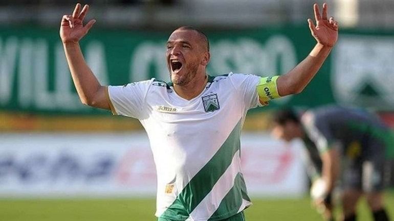 Un hombre con una camiseta de fútbol negra y roja, de pie en un campo. Tiene una expresión seria.