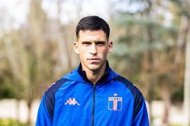 Un jugador de fútbol con una chaqueta azul con el logo del equipo; al aire libre.