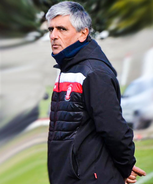 Un entrenador de fútbol con chaqueta mira hacia un lado, en un campo.
