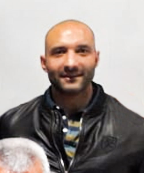 Hombre calvo, con barba oscura, vestido con una chaqueta negra sobre una camisa a rayas, sonriendo.