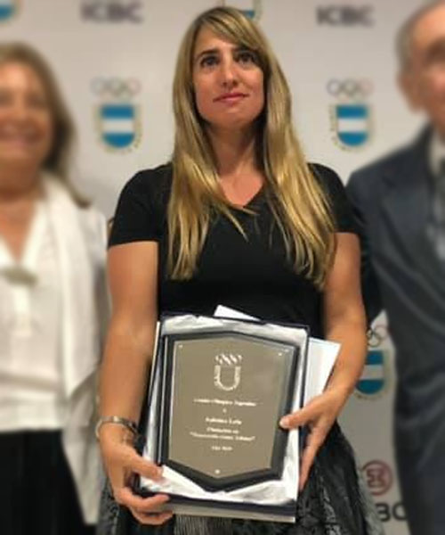 Mujer con cabello rubio sosteniendo una placa, vistiendo un top negro, en una ceremonia de premiación.