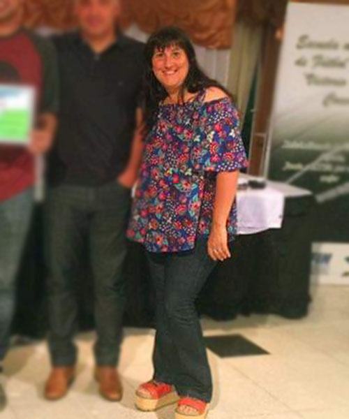 Mujer sonriendo con top floral, jeans y sandalias de plataforma; dos hombres borrosos en el fondo.