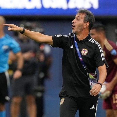 Entrenador de fútbol señalando con el brazo levantado; uniforme negro con insignia, fondo del estadio.