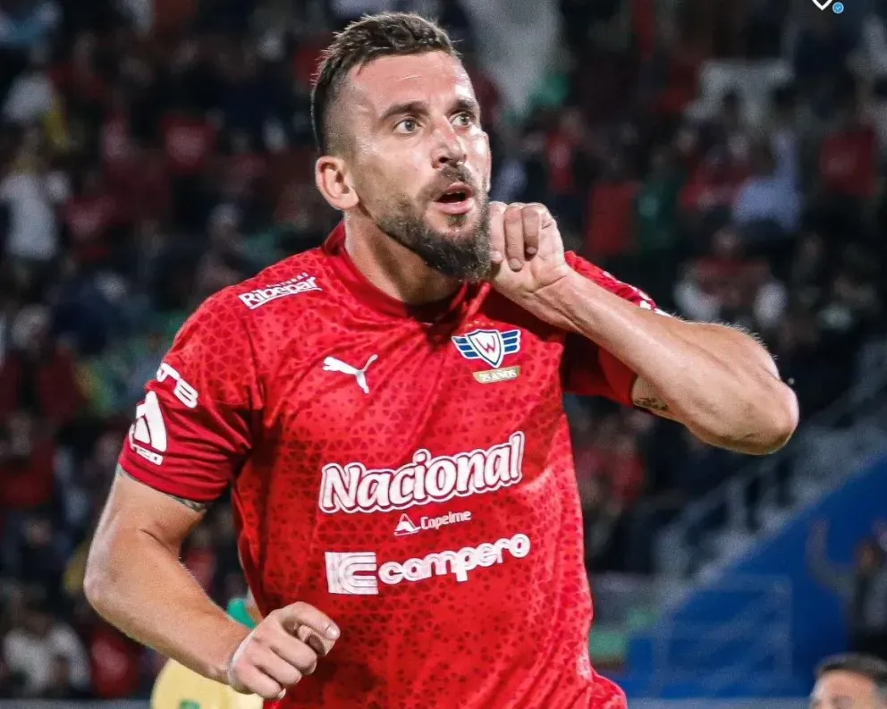 Un futbolista con camiseta roja celebra un gol levantando el brazo y con un estadio al fondo.