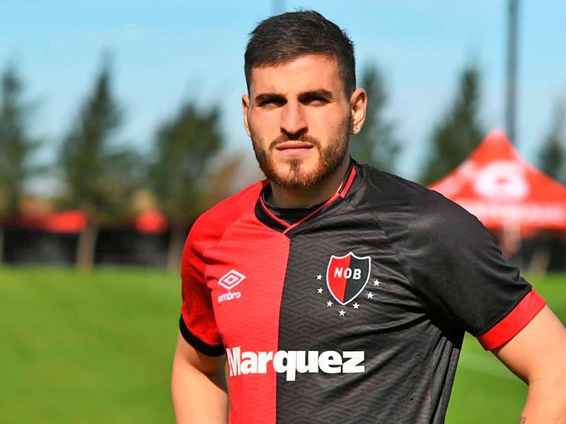 Un hombre con una camiseta de fútbol negra y roja, de pie en un campo. Tiene una expresión seria.