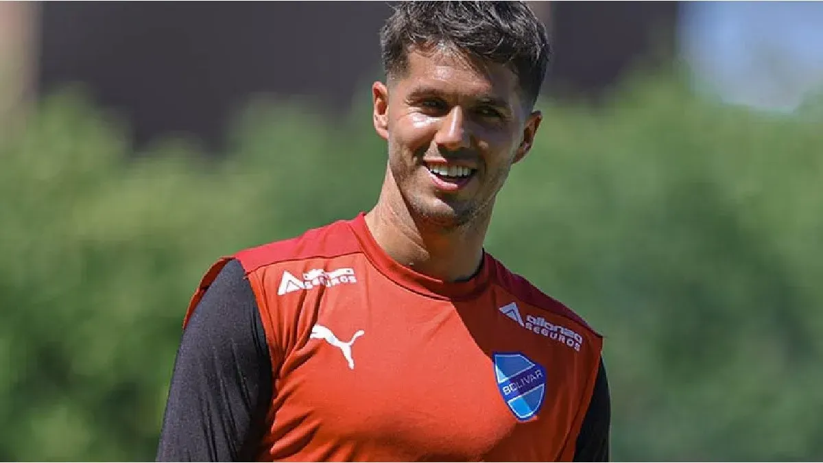 Jugador de fútbol sonriendo, vistiendo una camiseta de entrenamiento roja, al aire libre bajo la luz del día.