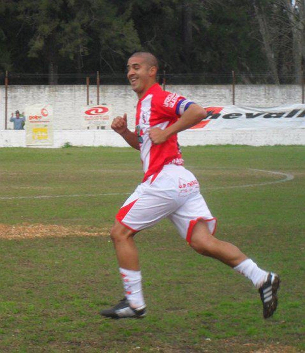 Jugador de fútbol con uniforme rojo y blanco, corriendo en un campo verde, sonriendo.