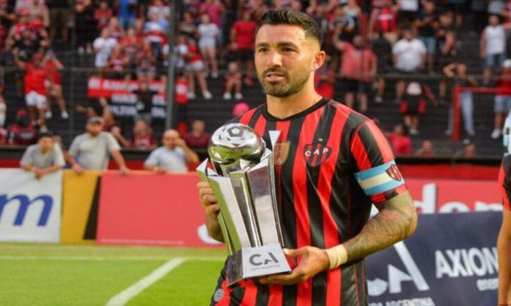 Jugador de fútbol con camiseta de rayas rojas y negras sosteniendo un trofeo en un campo, con multitud en el fondo.