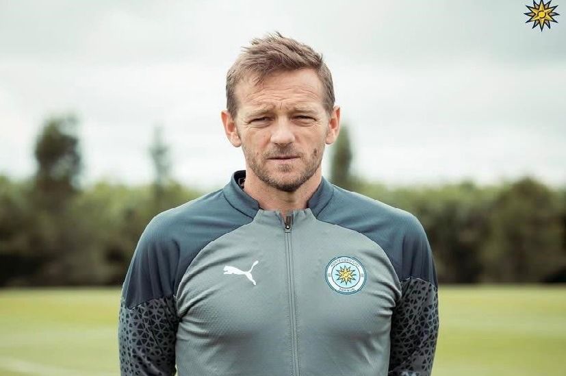 Un hombre con una chaqueta Puma gris con el logo del equipo, de pie afuera, con expresión neutral.