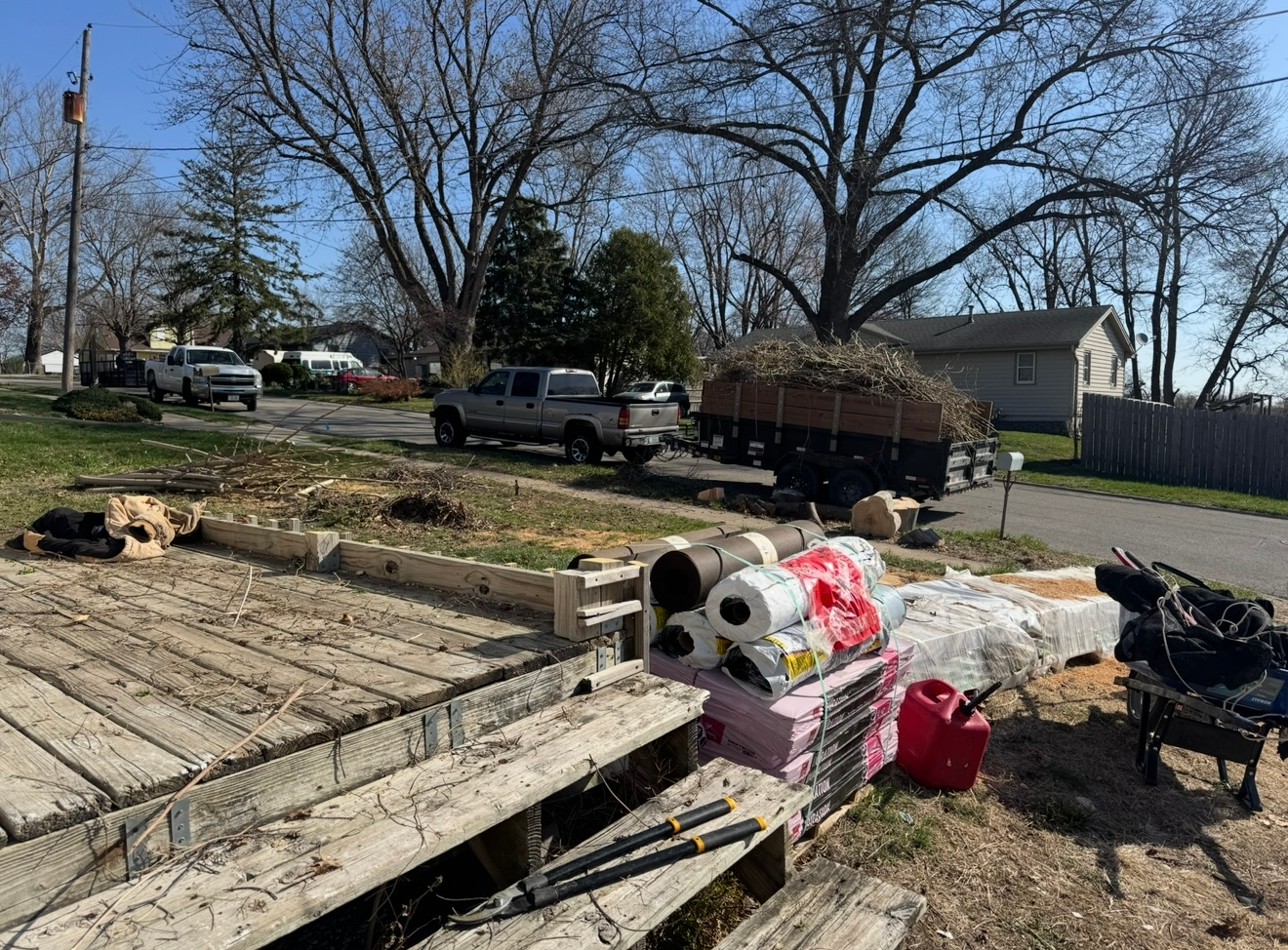 Debris pile with trash and a broken pallet beside a residential street and parked trucks under bare trees