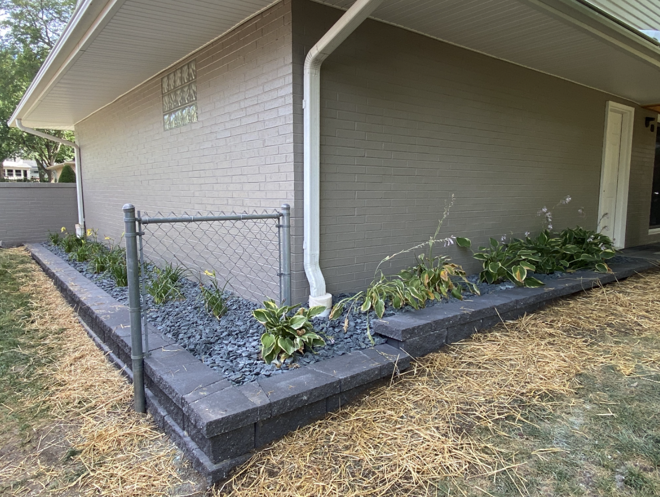 Corner of a beige house with a fenced garden bed and raised black edging, seen from the side.