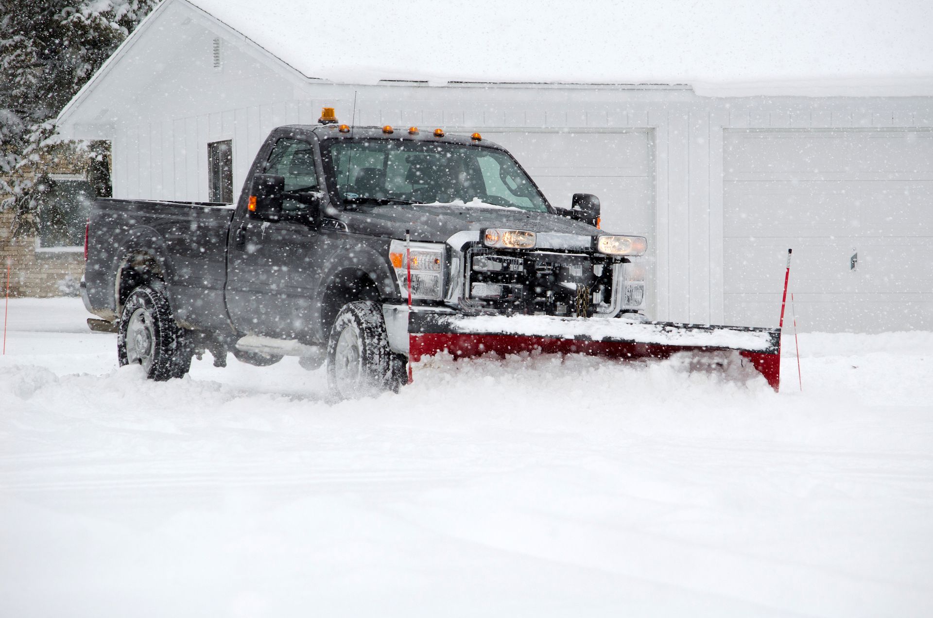 Black pickup truck with red snow plow clearing heavy snow in a driveway during a snowstorm