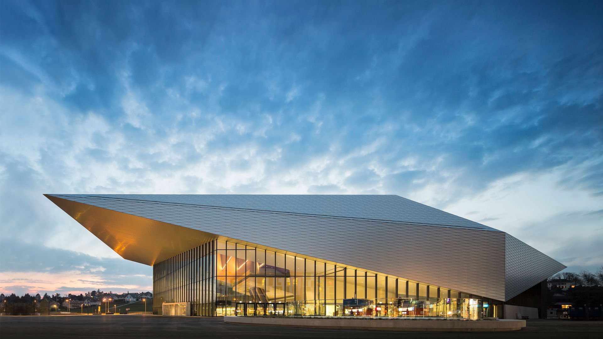 Modern building with angled roof and glass facade. Lit interior, cloudy sky.