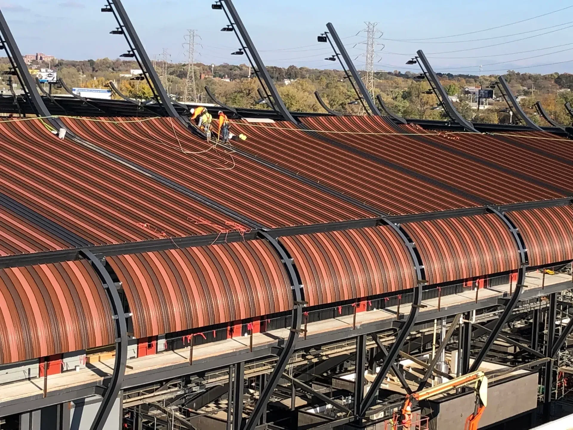 Construction workers on a stadium roof; brown metal panels, black support beams, and a blue sky in the background.