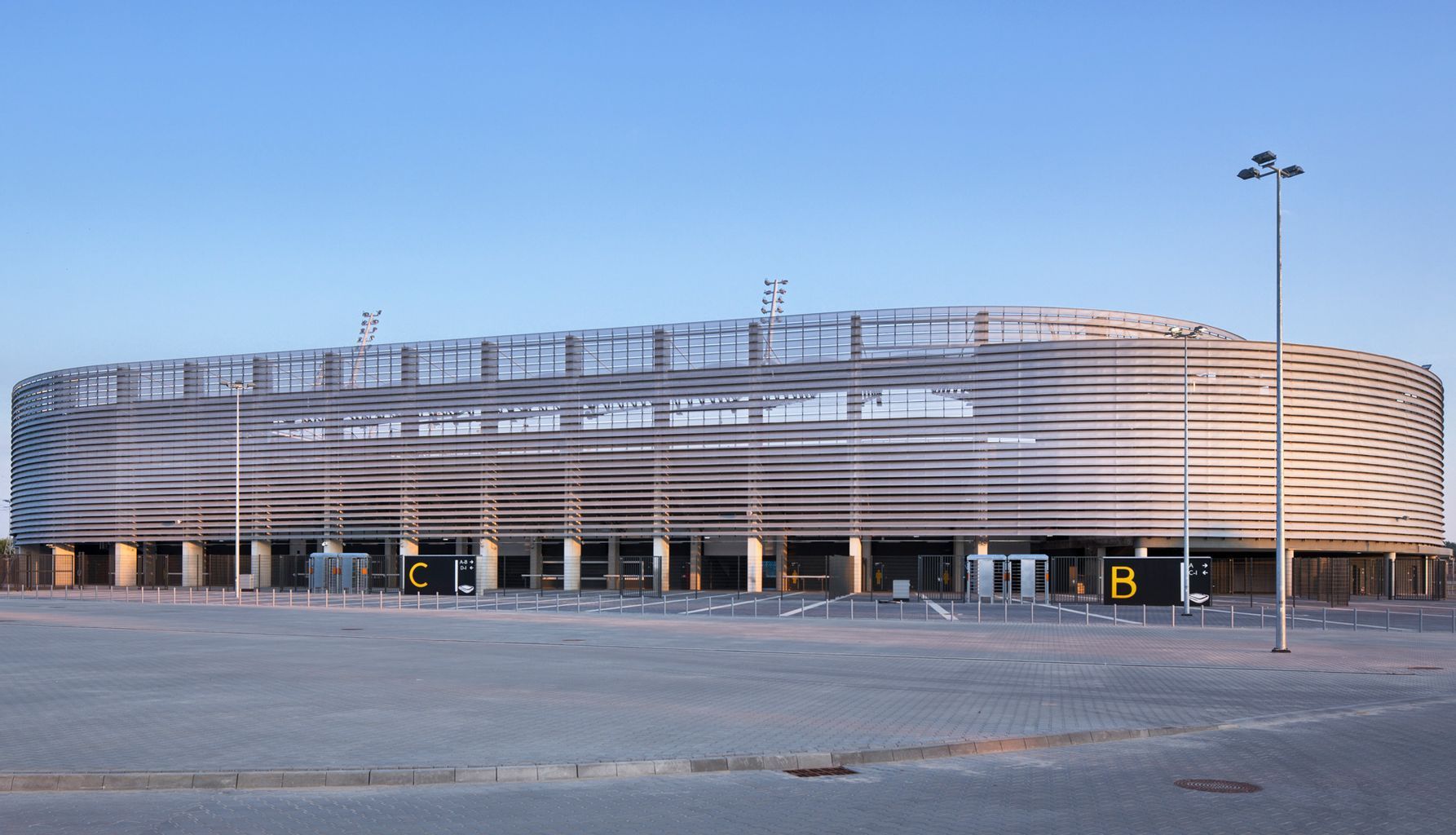 Modern stadium exterior with white, wave-like facade under a blue sky, with surrounding parking area.