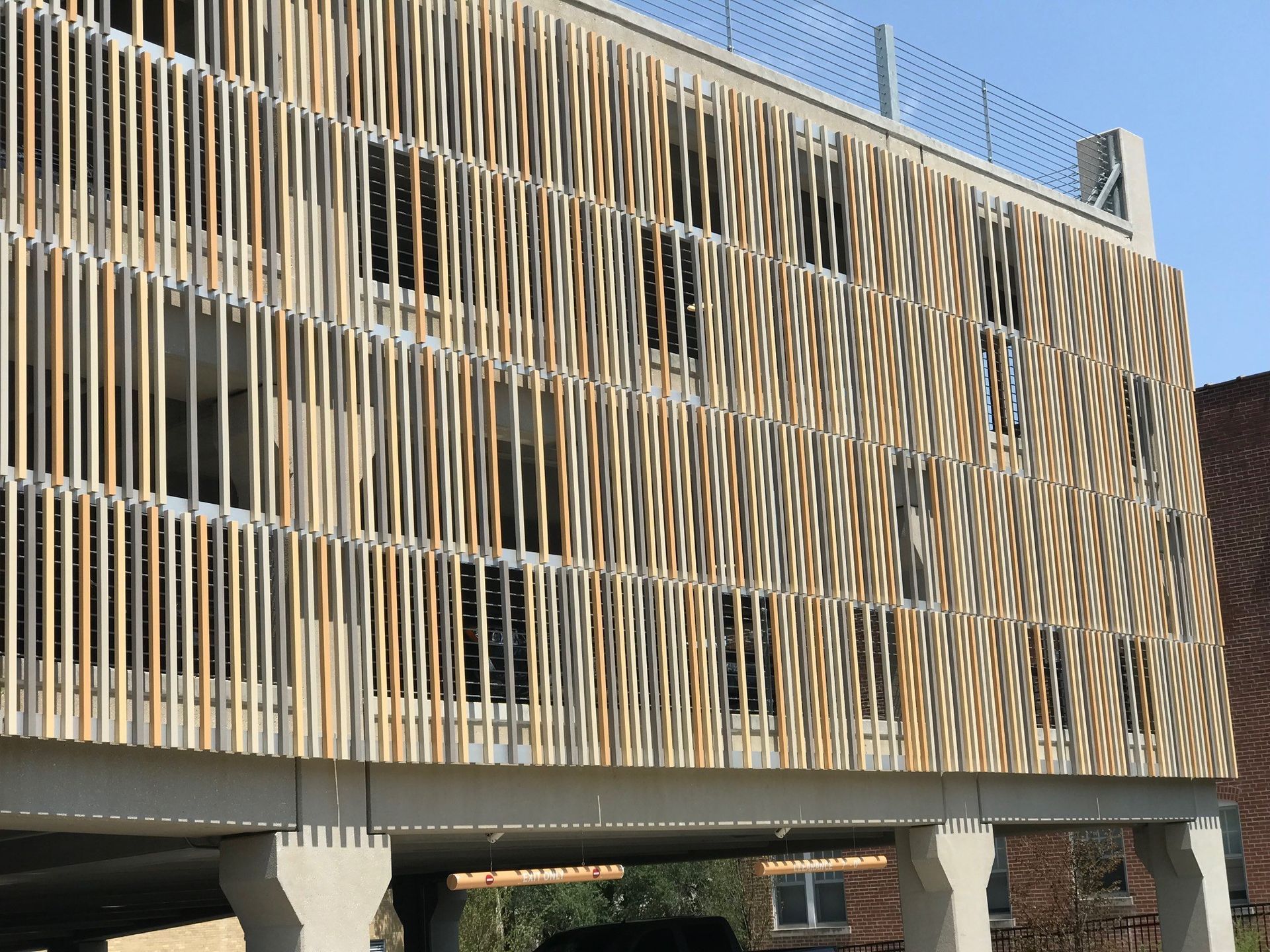 Wooden slat facade on a multi-story building. Concrete support columns below. Bright sunlight casts shadows.