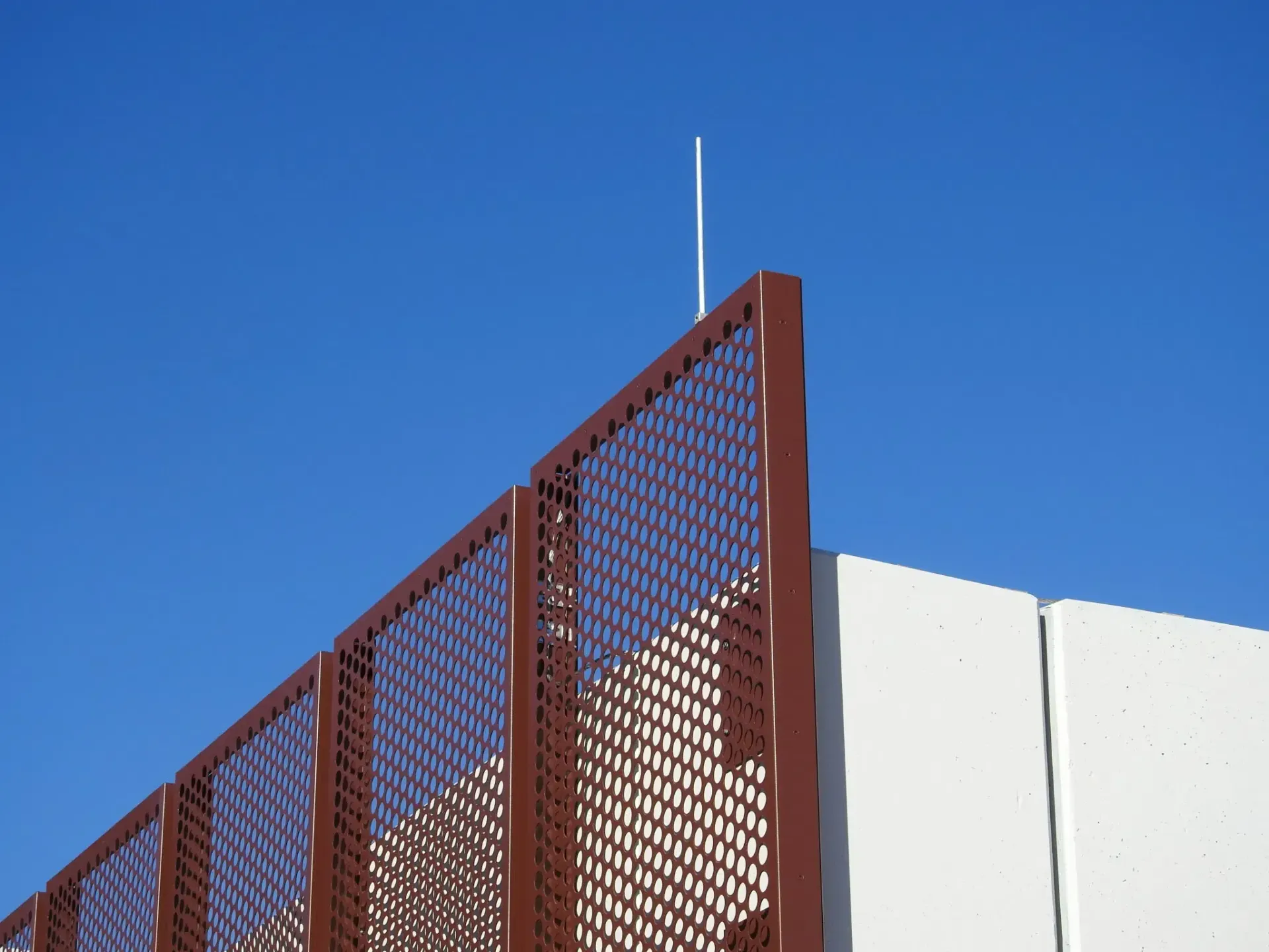 Brown metal fence with a grid pattern against a clear blue sky. A tall antenna sticks up.