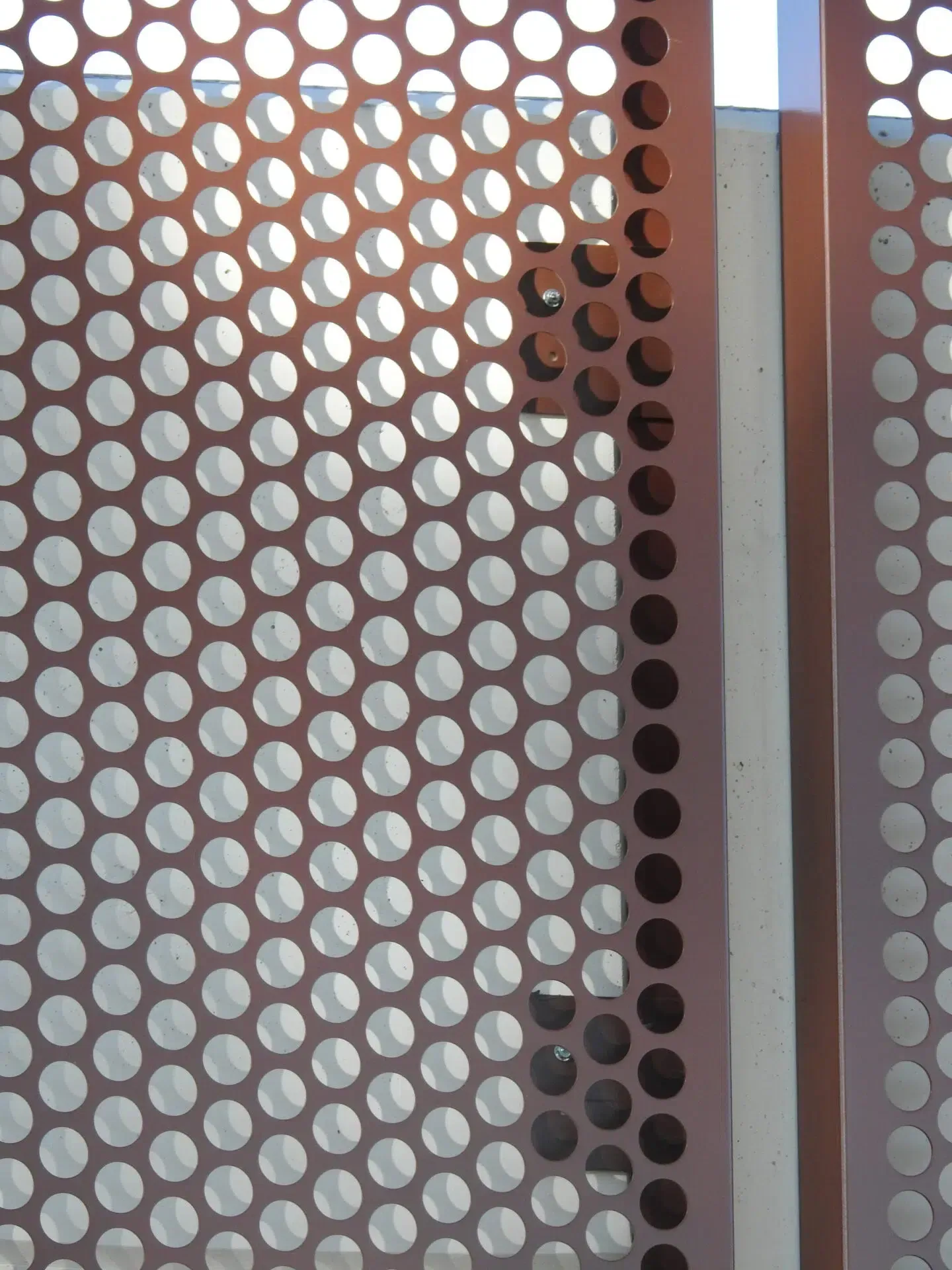 Perforated metal panel with circular holes, casting shadows. Brown and silver.