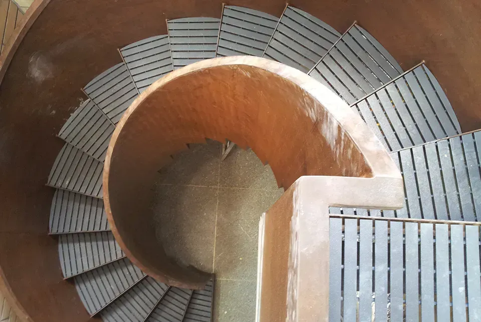 Spiral staircase, top-down view. Wooden steps lead down into a circular structure. Earth-tone walls.
