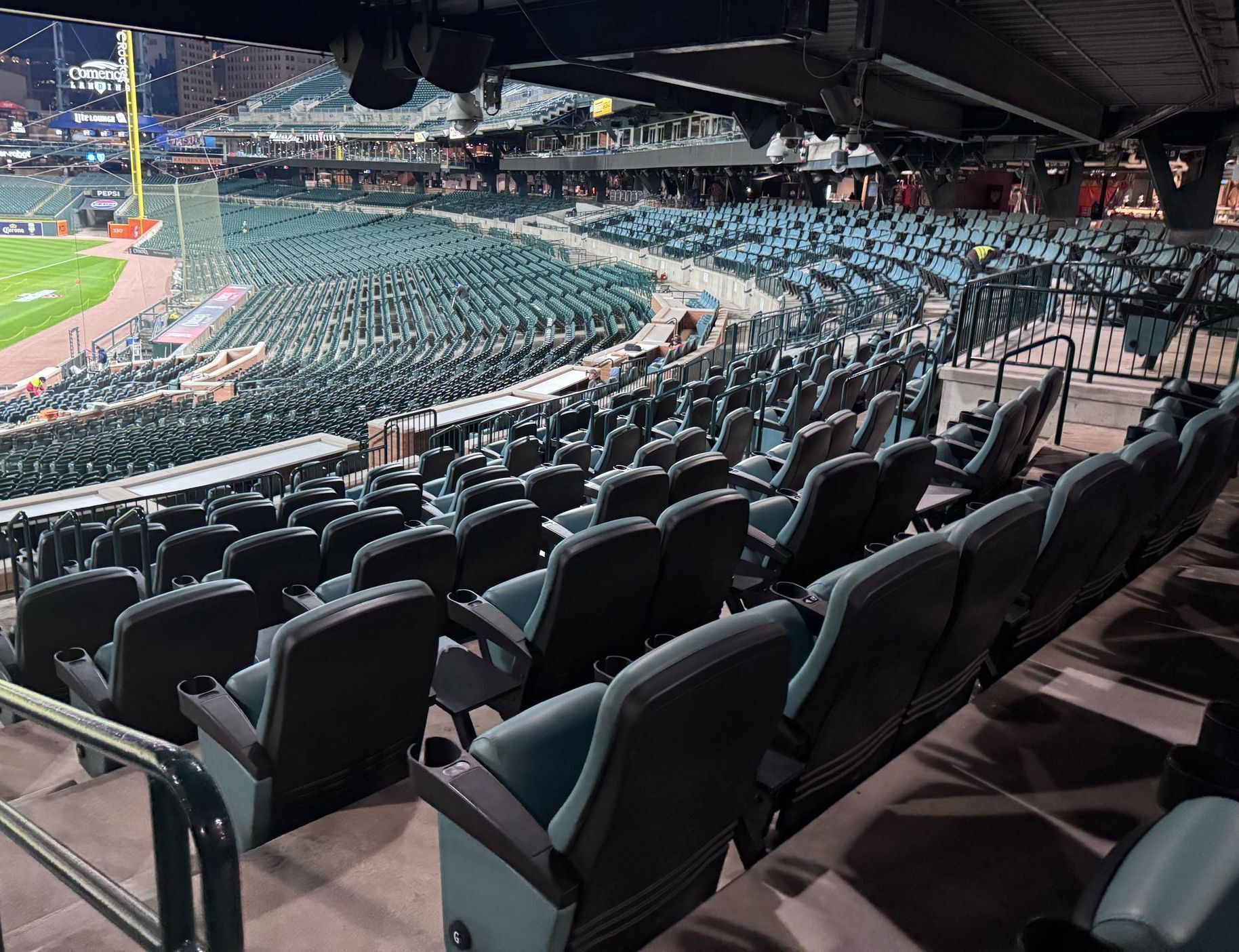 Empty baseball stadium seating, teal chairs, field visible, night.