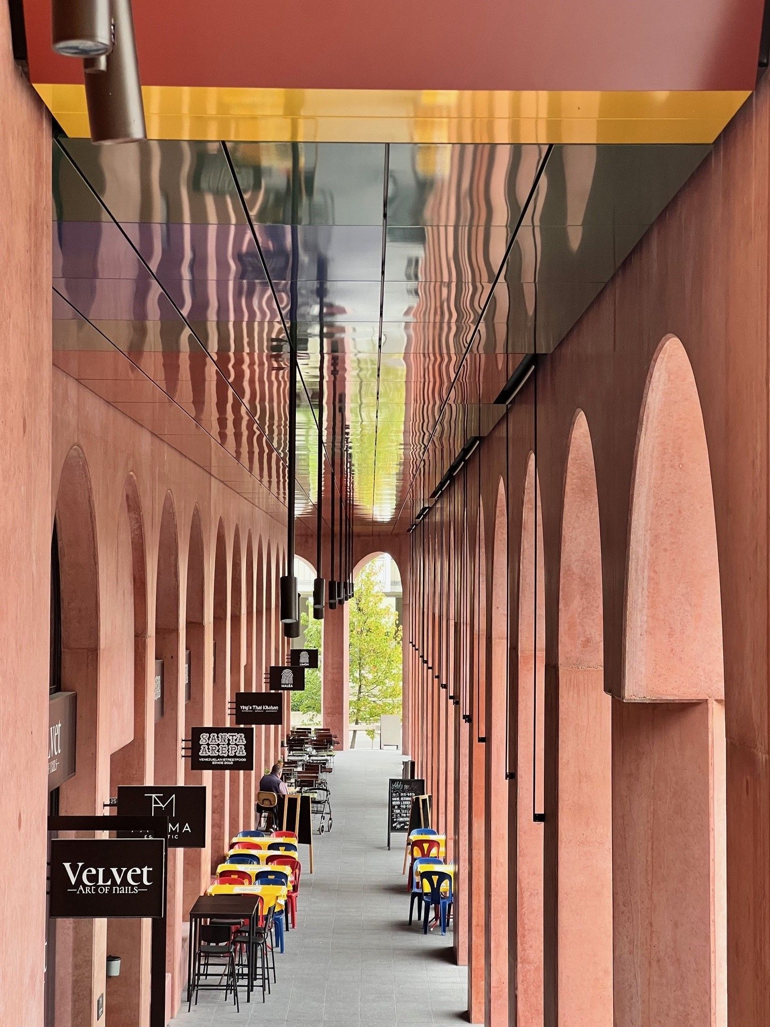 Arched walkway lined with outdoor tables and hanging signs. Red walls, reflective ceiling, sunny day.