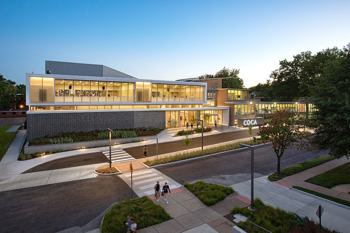 Modern building with glass walls, brick facade, and pedestrian walkway. Two people walk. Dusk setting.