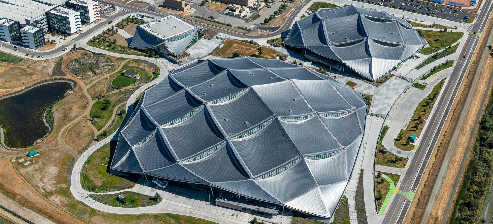 Aerial view of three modern, geometric buildings with patterned roofs, situated near roadways and a small pond.