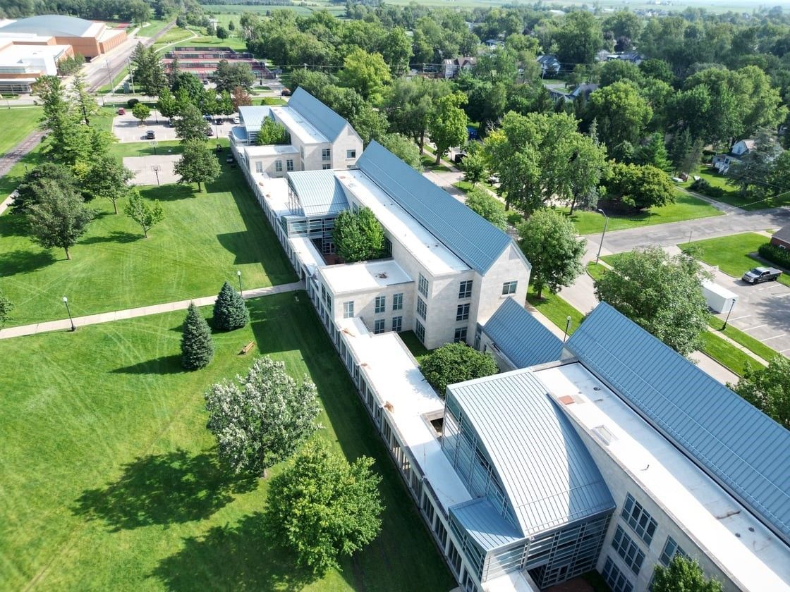 Aerial view of a long, light-colored building with a blue roof, surrounded by green grass and trees.