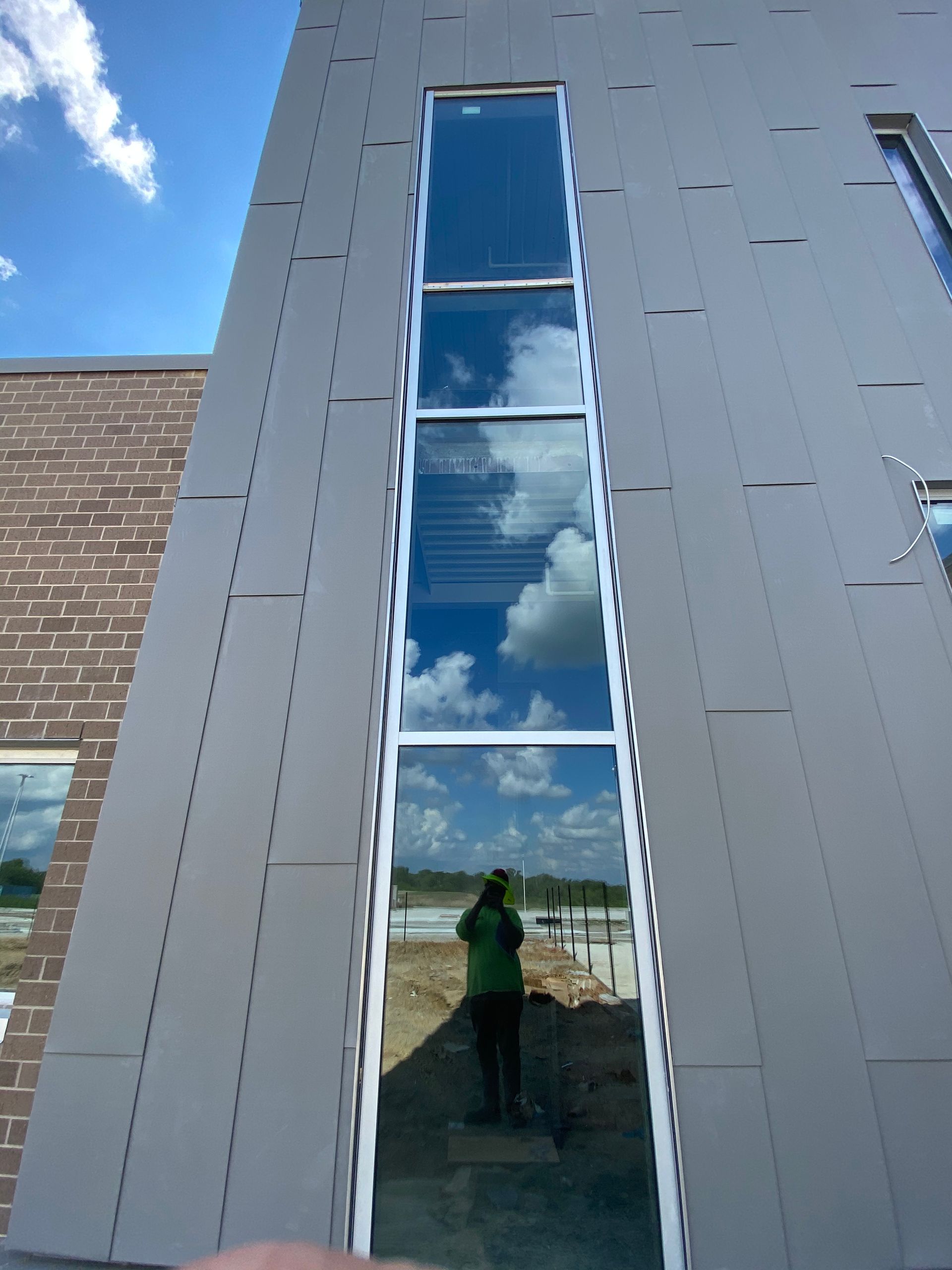Tall windows reflecting a person in green shirt, clouds, and a landscape. Gray building exterior and blue sky.