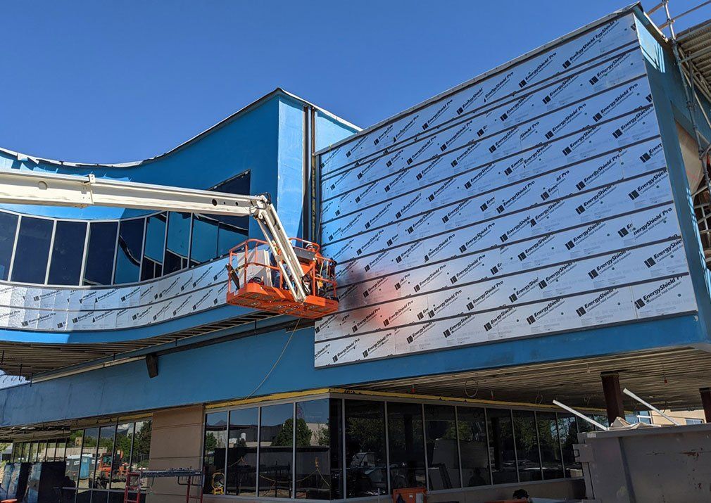 Construction workers on a lift installing silver panels on a light blue building under a clear sky.