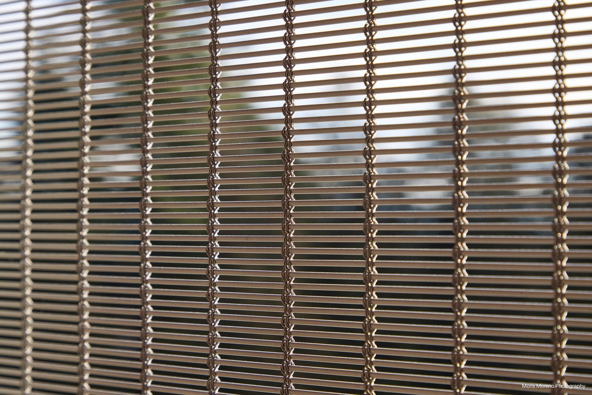 Close-up of a bronze-colored metal mesh curtain, with a blurry background of trees visible.