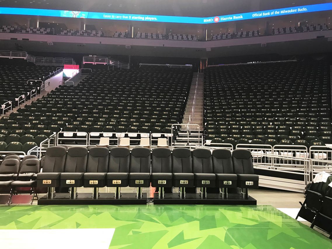 Empty arena seating with dark seats, and a row of black chairs on a green surface.