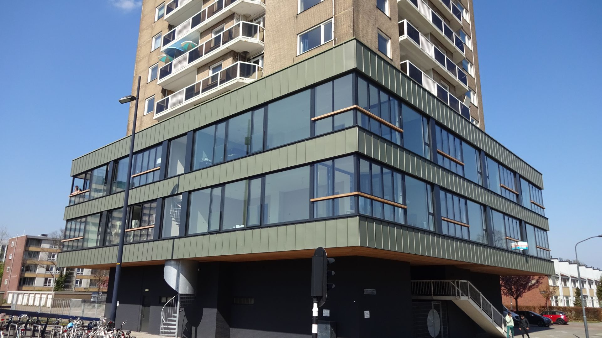 Multi-story building with glass windows, balconies, and a dark base. Blue sky overhead.