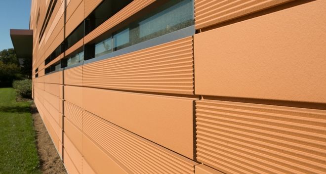 Exterior of a modern building with terracotta-colored panels, some with ribbed texture, beside a green lawn.