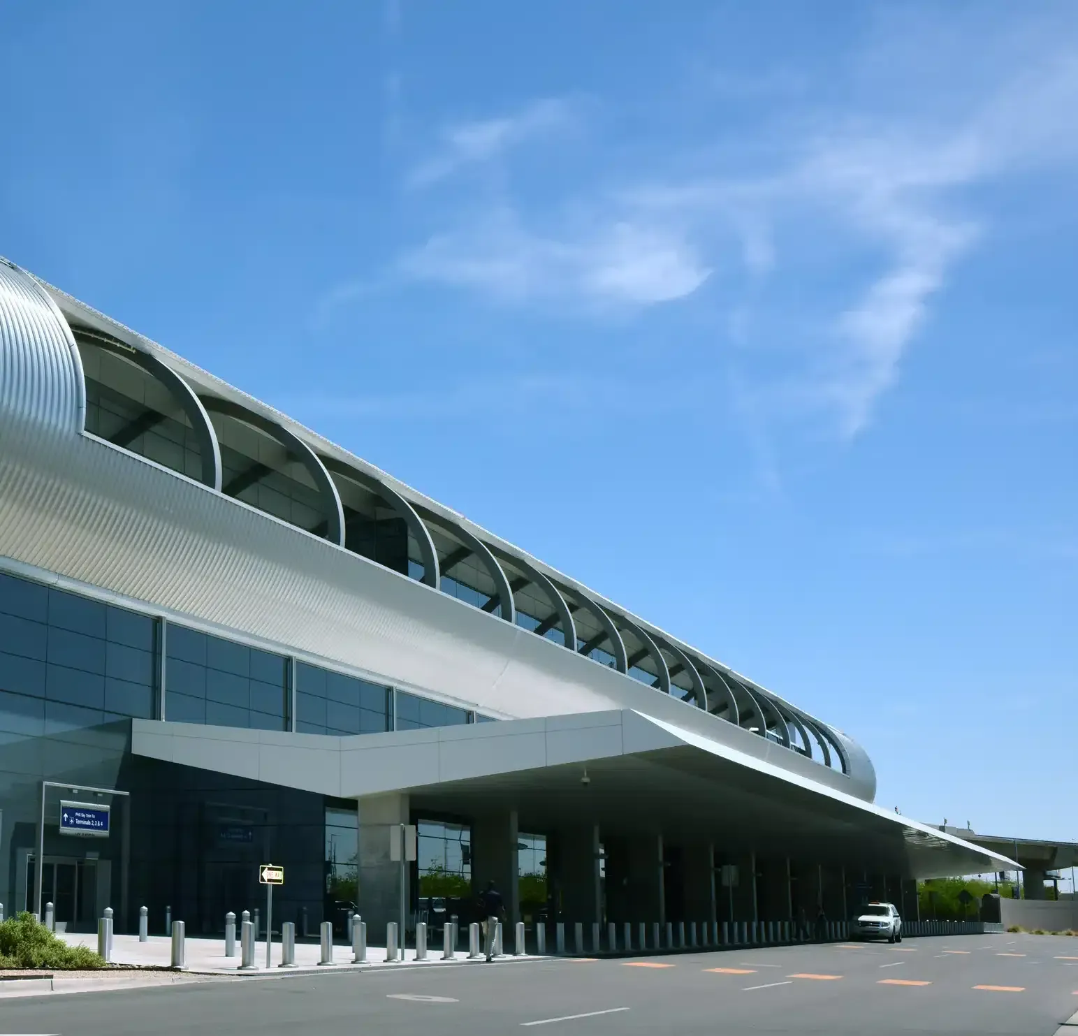 Exterior view of a modern airport terminal on a sunny day. Blue sky, metallic facade, and covered entryway.