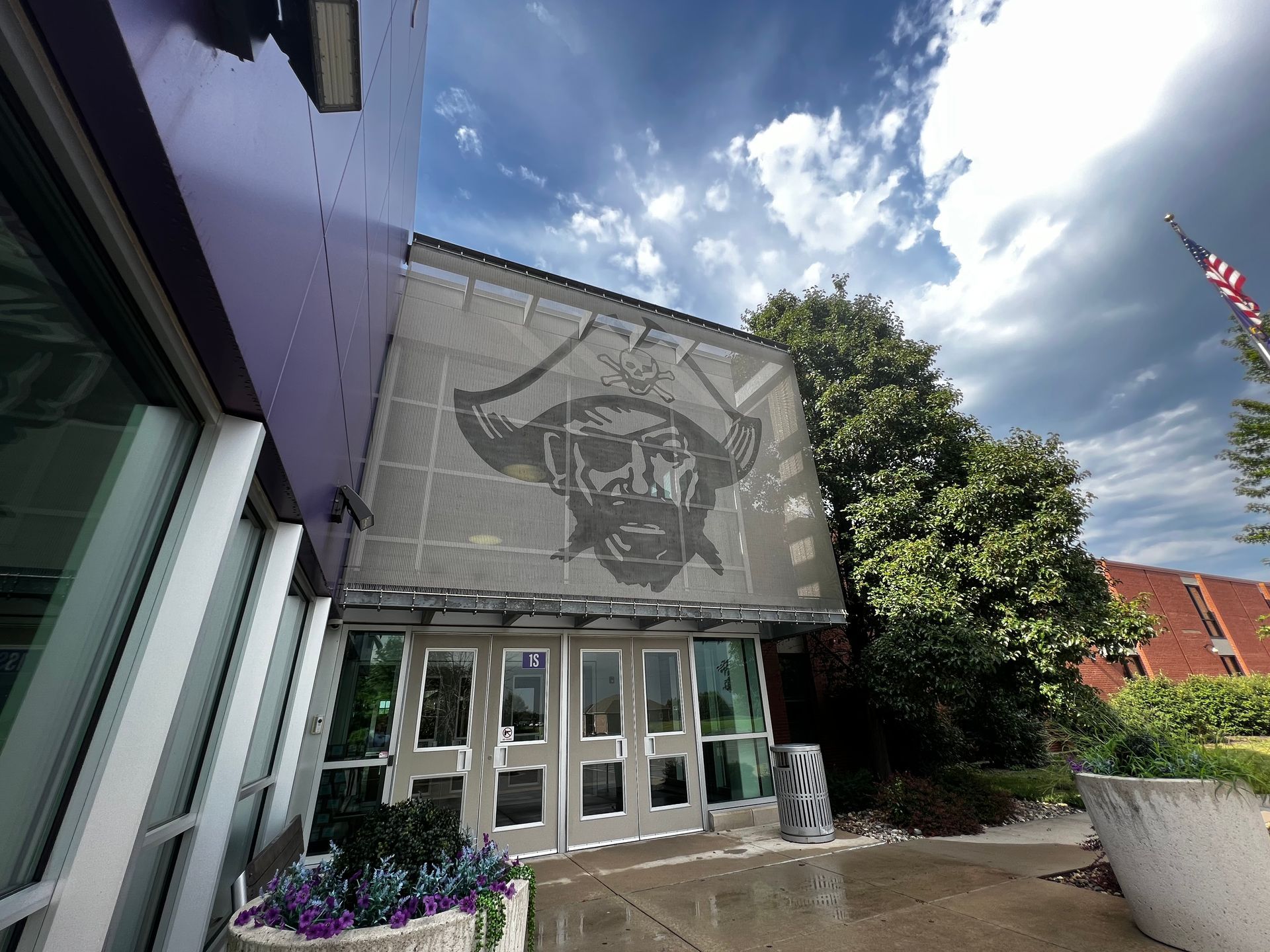 Entrance to a building with a pirate logo above doors. Purple wall, cloudy sky, American flag.